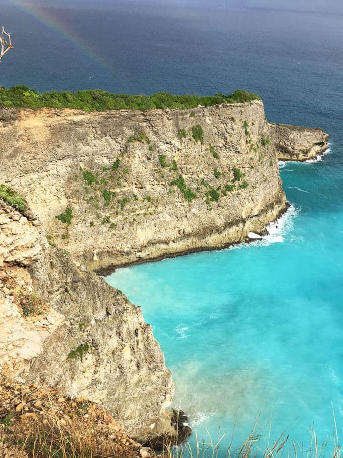 Sheer limestone cliff above turquoise water, with a rainbow out at sea