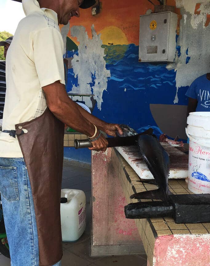 A fisherman in an apron cuts up a large fish with a knife on a tiled counter