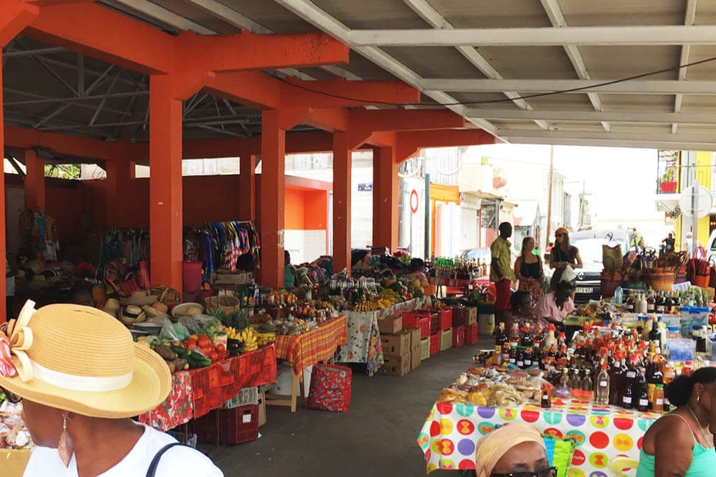 Inside the Grand-Bourg market hall: stalls of fruit, vegetables and bottles of punch between orange pillars