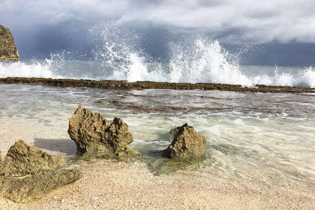 Waves breaking on a reef just below the surface, in front of a sandy beach and a few rocks